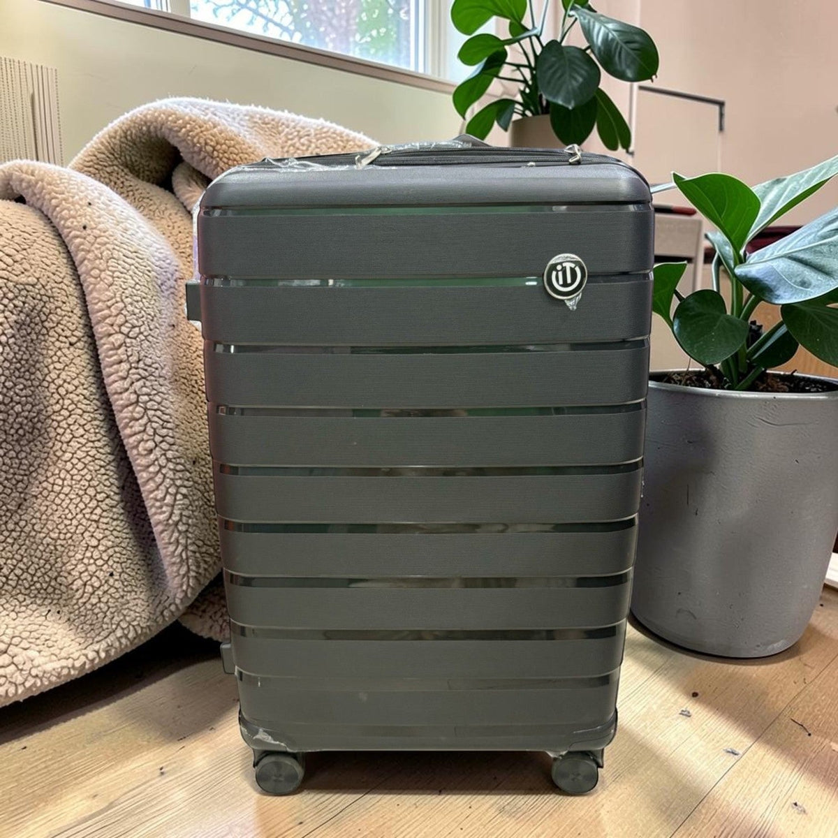 Fixed Price Shop grey hard shell spinner suitcase with horizontal ribbed design and IT logo on a wooden floor.